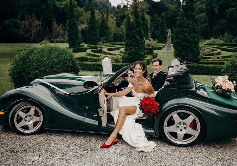 Bride and groom in sports car, bride is wearing red shoes and holding a bouquet of red roses